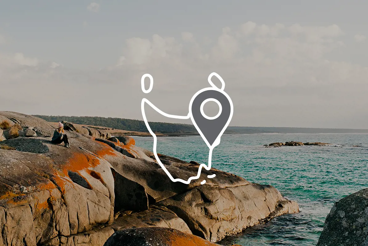 A person sitting on rocks next to the ocean. Overlaid is an icon of the outline of Tasmania with a map pin.