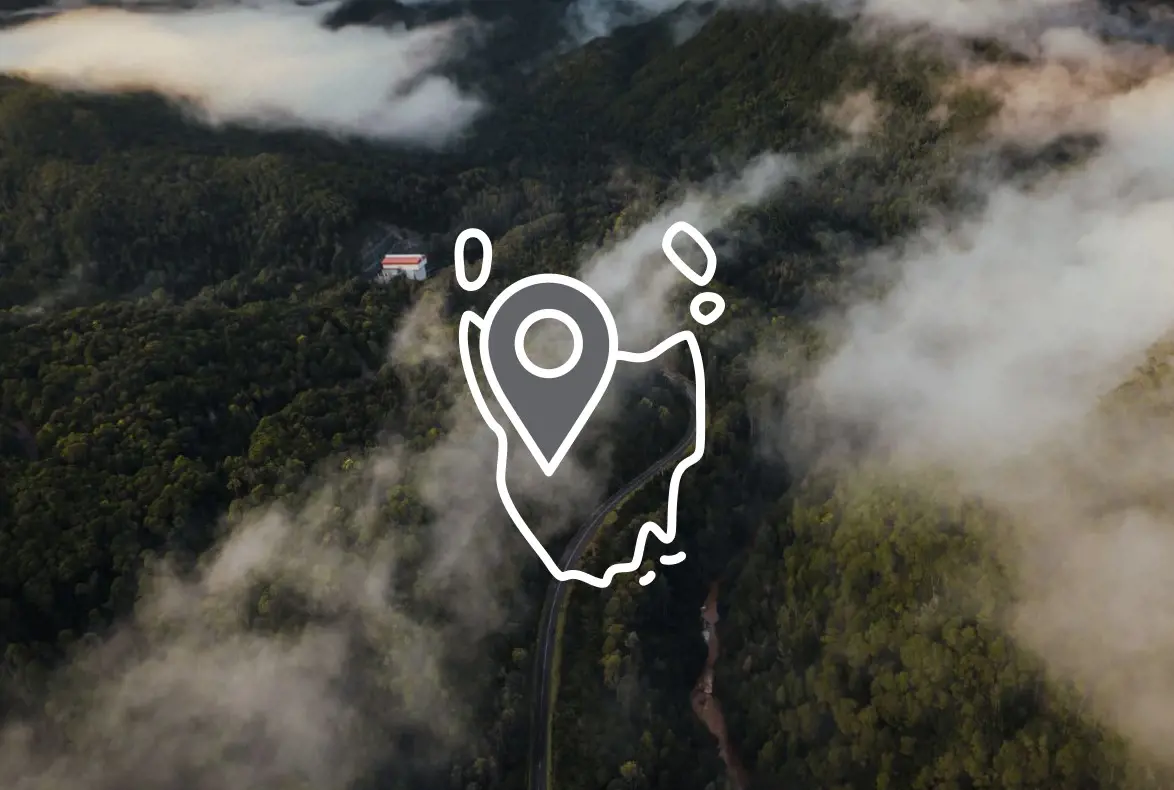 An aerial view through wispy clouds of dense bush, with a small road running through. Overlaid is an icon of the outline of Tasmania with a map pin.