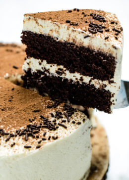 Chocolate coffee cake with a slice of cake being lifted up. The cake is coated in mascarpone frosting, with a middle layer of frosting, and decorated with chocolate sprinkles and a dusting of cocoa powder.