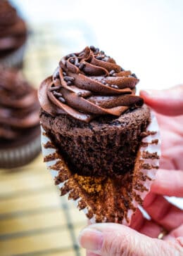 Two hands pulling the paper liner off of a gluten free chocolate cupcake with more cupcakes in the background.