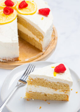 A lemon cake topped with lemon slices, sprinkles, and raspberries on a wooden cake stand, with a slice in front of the cake on a plate with a fork.