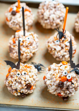 Halloween popcorn balls on a sheet pan surrounded by orange sprinkles and spiders, topped with bat toothpicks.