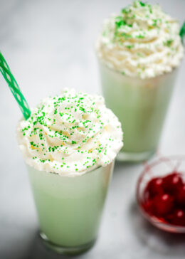 A tall glass with shamrock shake topped with whipped cream and sprinkles, next to a bowl of maraschino cherries. There is another shake in the background, and they both have straws.