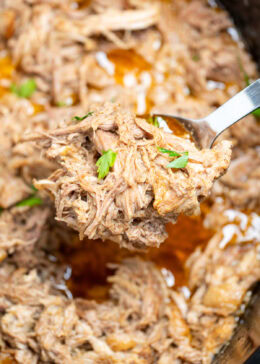 A slow cooker full of shredded pork shoulder in broth and drippings, with a spoon lifting some up.