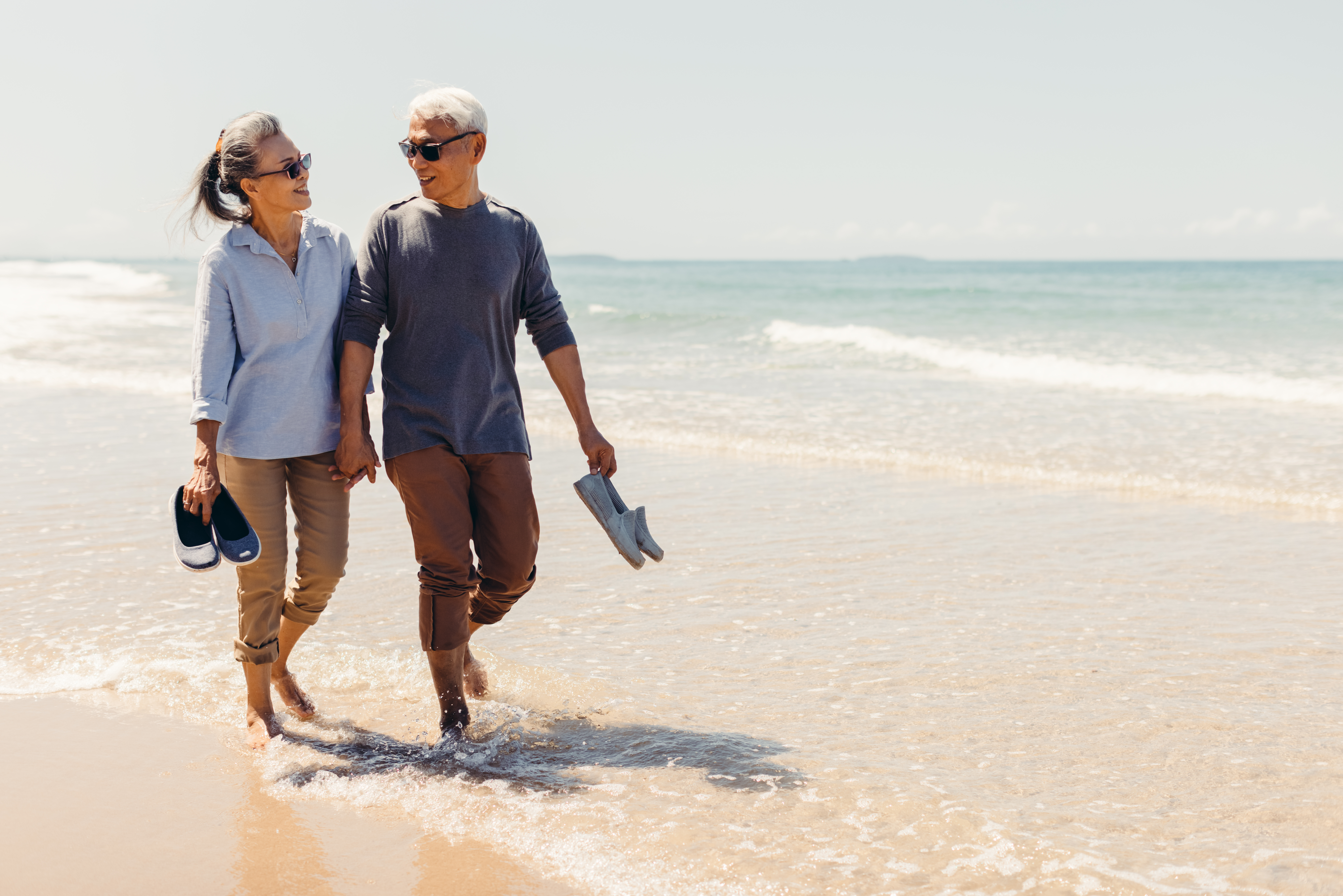 A couple walking hand-in-hand along the beach, smiling at each other, barefoot with their shoes in hand, as gentle waves lap at their feet.