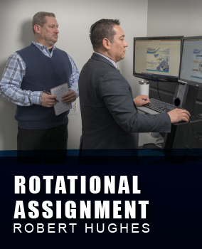 Two men in an office setting, with one standing and typing at a computer workstation with dual monitors, while the other holds a notepad and observes. Both men are engaged in a work-related task. The banner below them says 'Rotational assignment Robert Hughes'