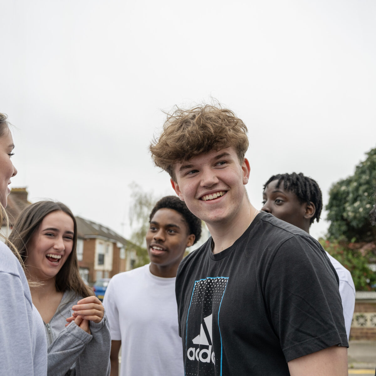 A group of five young people is standing outside, engaged in conversation and laughing. The central figure is a young man with curly hair wearing a black Adidas t-shirt. They all appear to be enjoying each other's company, with a backdrop of houses and trees under an overcast sky. The atmosphere is casual and friendly.