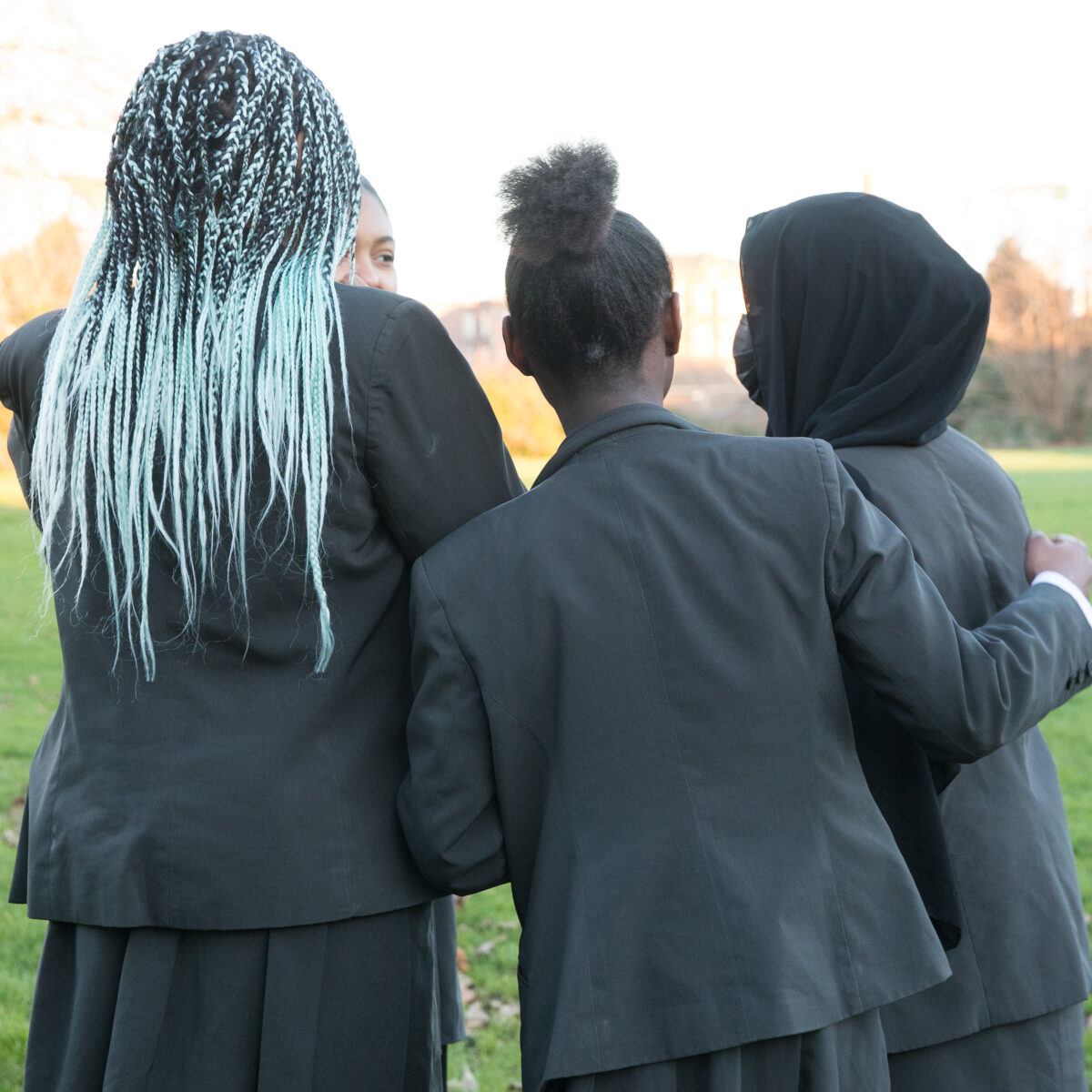 Four individuals in school uniforms, photographed from behind, are standing closely together outdoors. One person's hair is braided with light blue extensions. They appear to be in a grassy area.