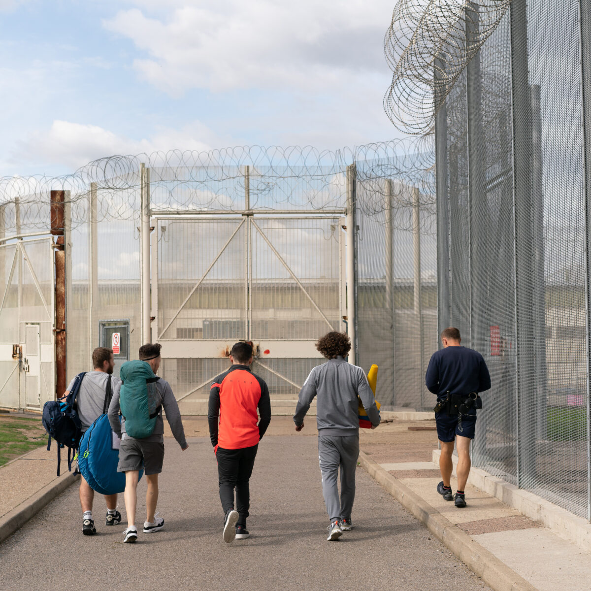 Five men walk towards a secured gate with barbed wire fencing, carrying bags and equipment under a cloudy sky.