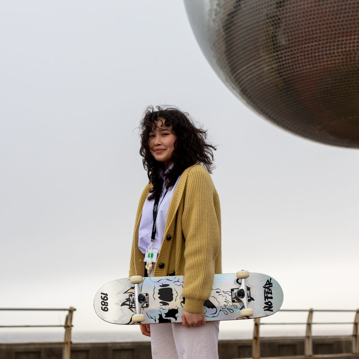 A young person holding a skateboarding. She is wearing a yellow cardigan and is outside standing in front of a circular sculpture.