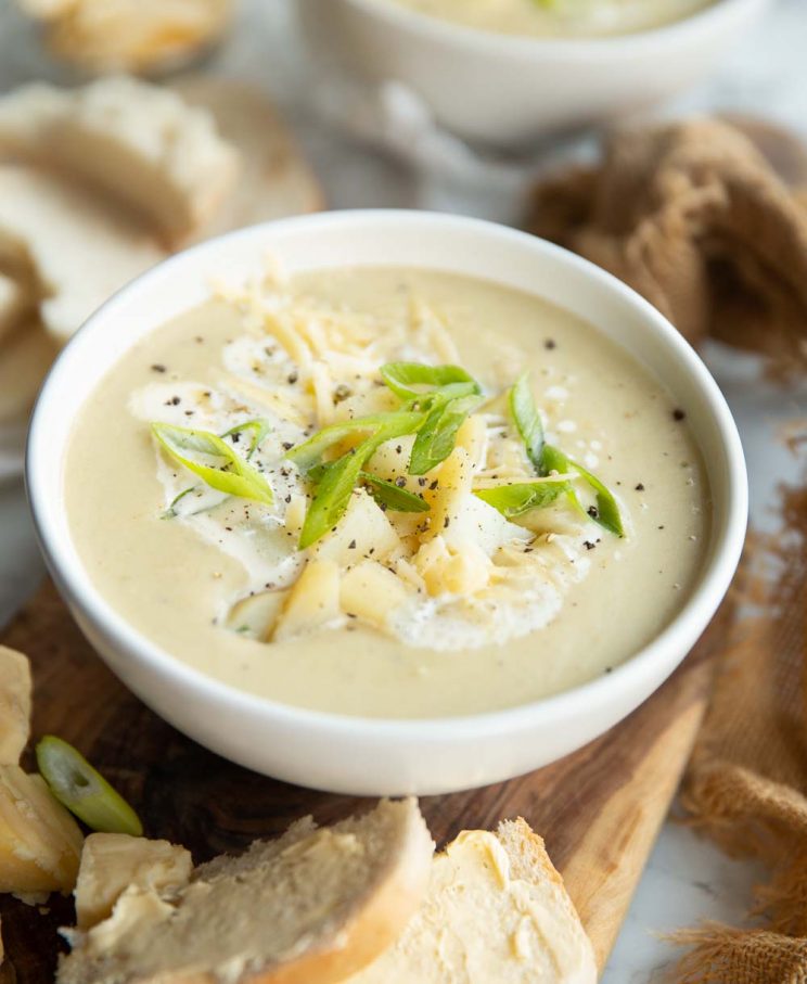 soup in a white bowl on chopping board surrounded by bread garnished with cheese, potato and spring onion