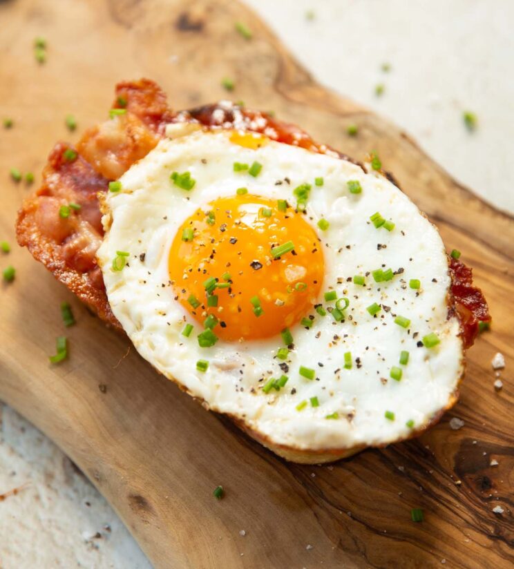 close up shot of egg and bacon on toast on wooden board garnished with chives