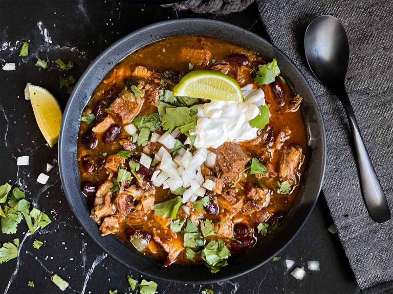 Smoked pork chili garnished with cilantro, onion, lime, sour cream, and onion in a dark bowl.