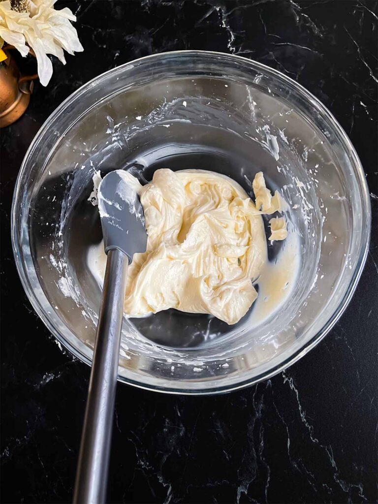 Softened cream cheese in a glass mixing bowl on a dark surface.