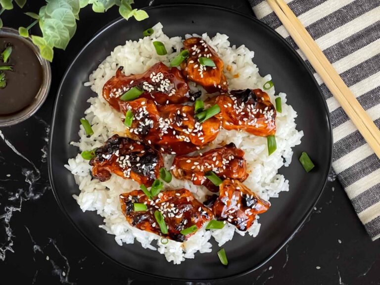 Bourbon chicken on a bed of white rice on a dark plate, garnished with sesame seeds and green onion.