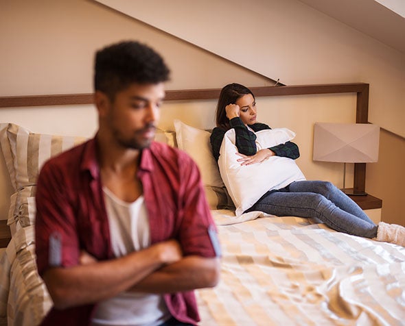 A couple sits on a bed turned away from each other, disgruntled.