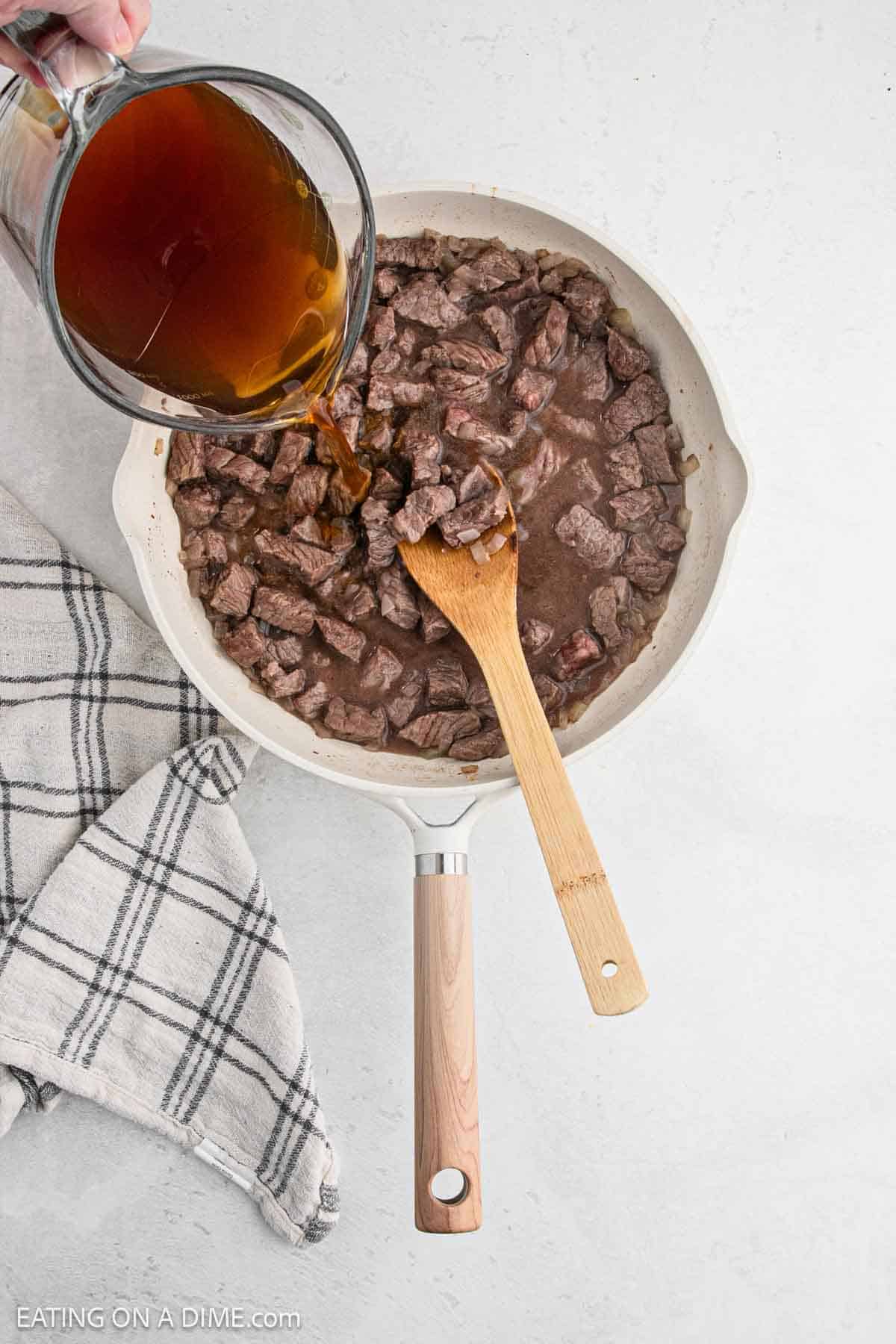 Pouring broth in a skillet with cooked beef tips with a wooden spoon