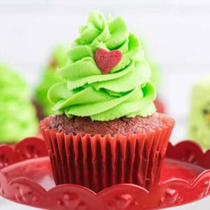 A chocolate cupcake with bright green frosting, whimsically resembling a Grinch-style Christmas tree, is topped with a small red heart. It sits on a decorative red plate, while other similar Grinch-themed cupcakes are blurred in the background.