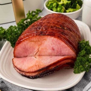 A glazed air fryer ham with a few slices cut is displayed on a white platter. The ham is garnished with parsley and accompanied by a bowl of broccoli in the background.