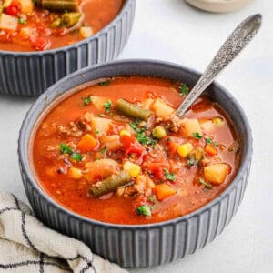 A bowl of hearty vegetable beef soup, reminiscent of a classic hamburger, with green beans, corn, potatoes, carrots, peas, and tomatoes in a rich broth. The textured bowl holds a resting spoon and sits beside a cloth napkin on a light surface—an inviting dish perfect for any recipe collection.