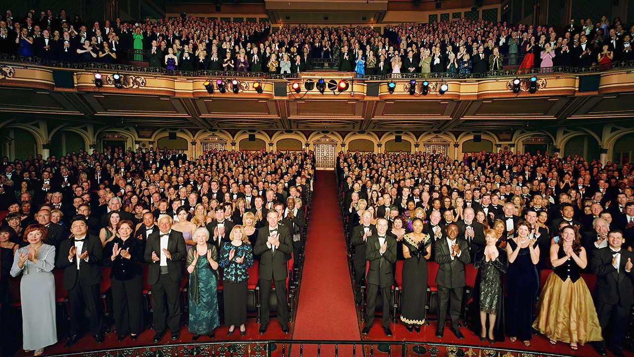 Theatre audience standing in formal attire, applauding.