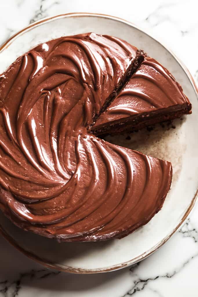 A top-down view of a whole devil's food cake with one slice partially removed, showcasing the rich, swirling chocolate frosting on top of the cake, set on a white plate with a gold rim, on a marble countertop.