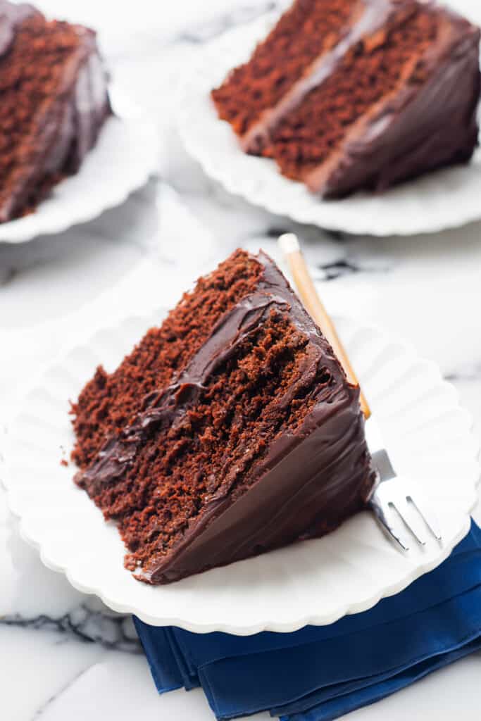 A slice of devil's food cake with glossy chocolate frosting on a white scalloped plate, with a fork on the side and a dark blue napkin underneath, set on a marble surface.