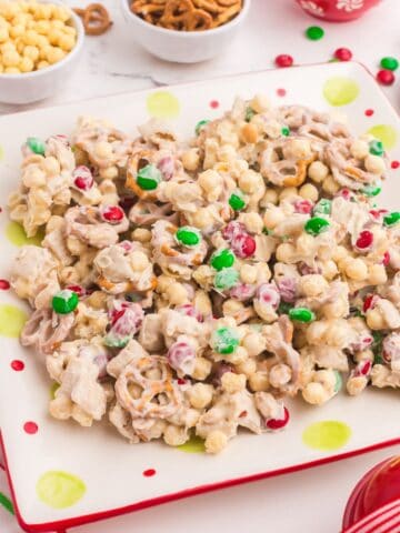 White trash candy (white chocolate chex mix candy) on a plate surrounded by small bowls of peanuts, cereal, pretzels, and Christmas colored M&Ms.