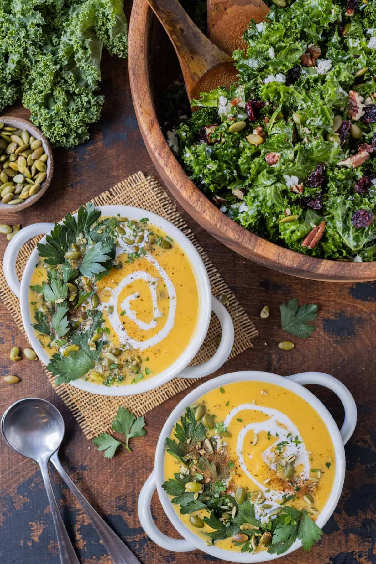 Two bowls of butternut squash soup with coconut milk drizzle and parsley leaves are next to a chopped kale salad and pumpkin seeds.