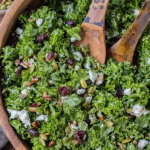 A chopped kale salad with cranberries, goat cheese, pecans, and pumpkin seeds in a wooden bowl with serving spoons for a Fall dinner.