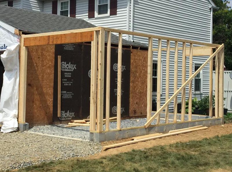 A partially constructed wooden frame structure is attached to the side of a white house in Madison, WI. The framing includes vertical and diagonal beams, and the ground is covered with gravel. Evident exterior renovations feature the house's white siding complemented by maroon shutters.