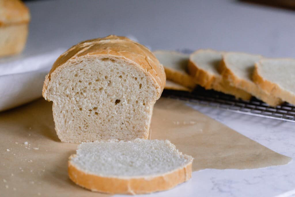 sourdough sandwich loaf with a slice of bread laying in front of the loaf on payment paper. More slices are in the background