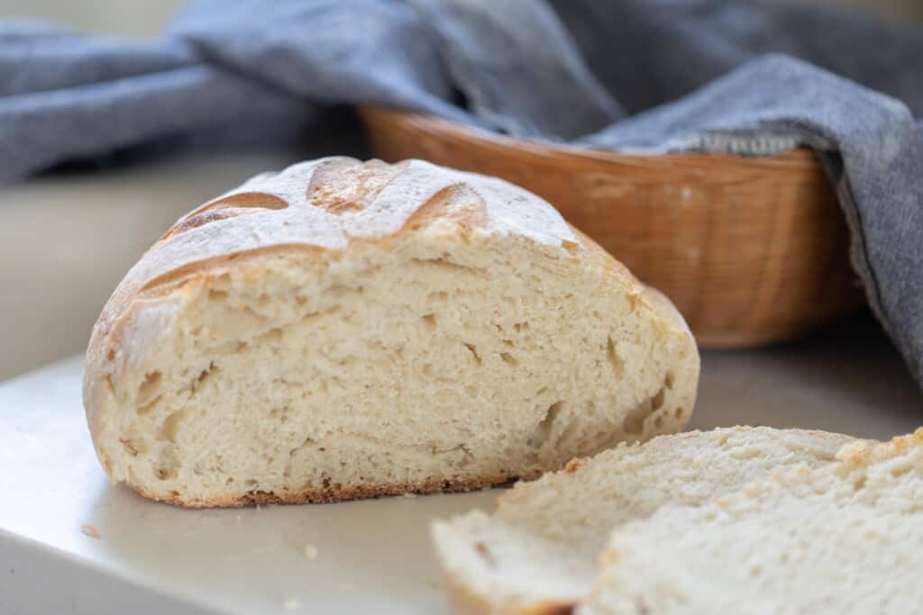 loaf of same day sourdough bread boule on a white countertop with two slices laying beside it. In the background there is a basket lined with a blue towel