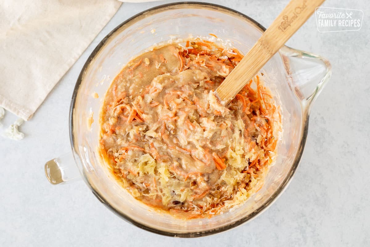 Glass mixing bowl with wooden spoon stirring in the carrots, coconut, pineapple and walnuts.