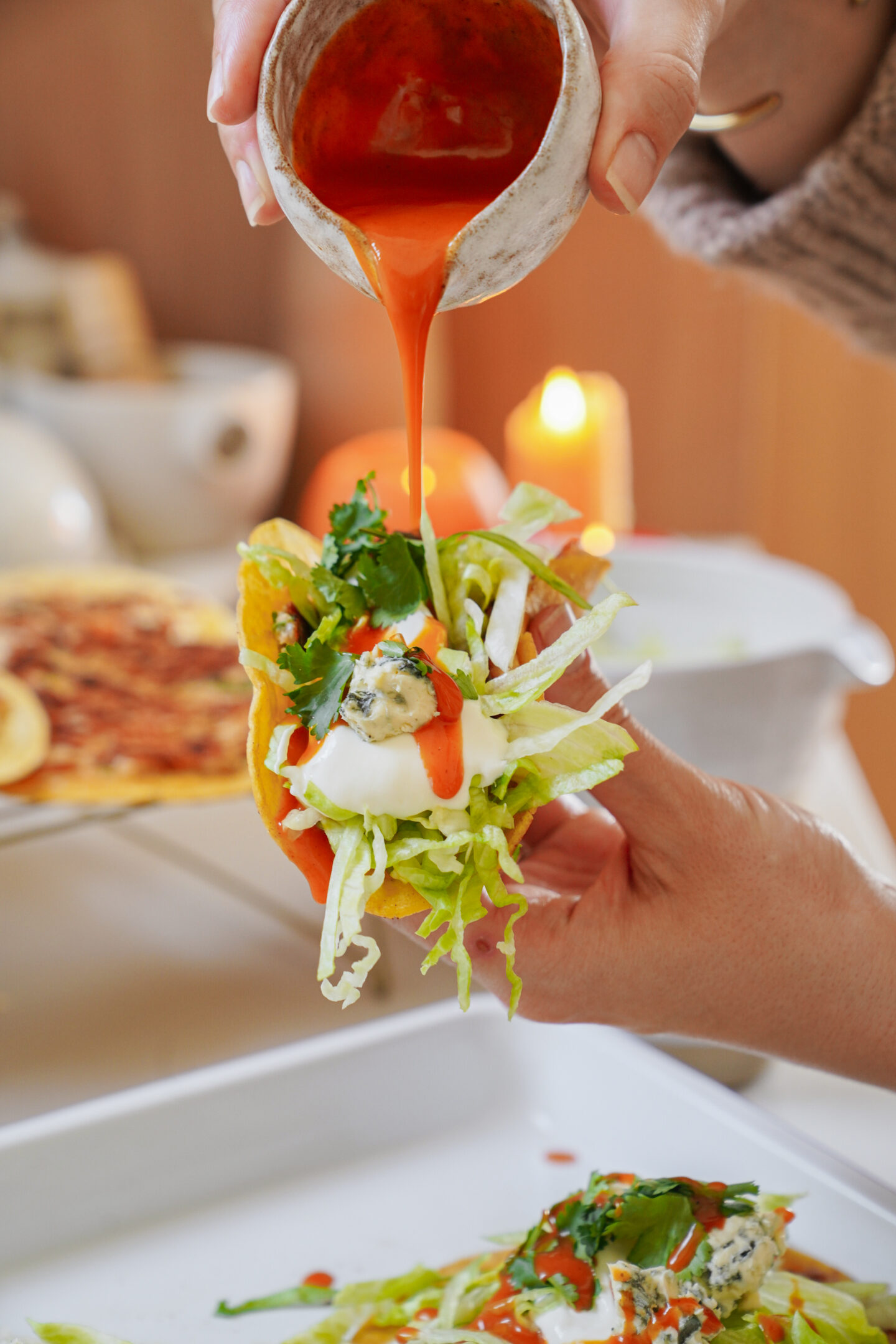 A person holds a taco filled with lettuce, sour cream, and other toppings. Another hand is pouring red sauce onto the taco from a small cup. A lit candle and a plate of food are blurred in the background.