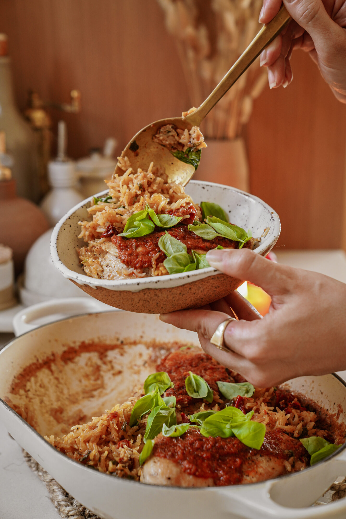 Person serving a portion of baked chicken topped with tomato sauce and fresh basil onto a plate. The dish is garnished with basil and placed in a rectangular baking pan on a wooden table setting. Hands are seen holding the plate and serving with a spoon.