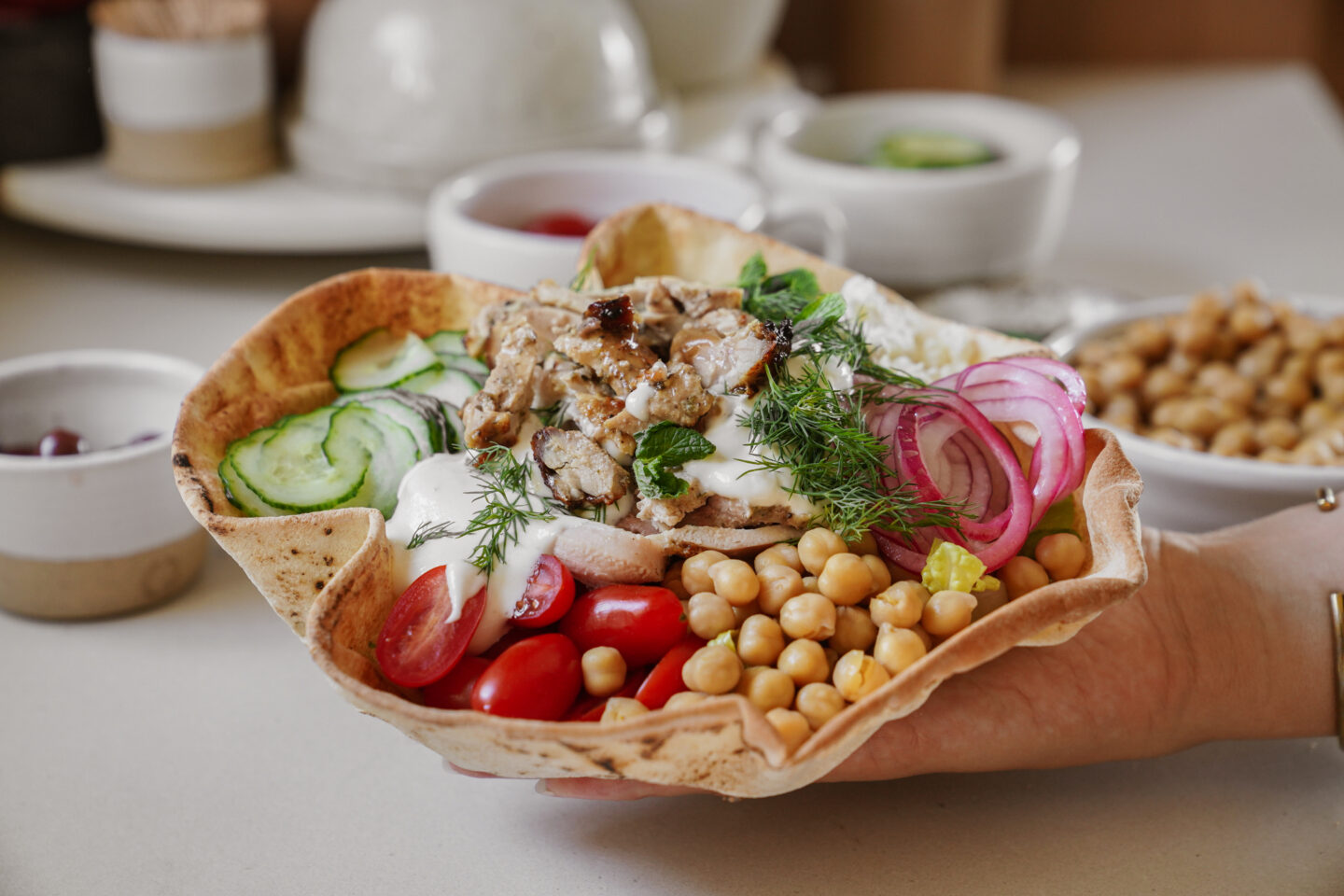 A hand holds a pita bowl filled with grilled meat, sliced cucumbers, red onions, cherry tomatoes, chickpeas, fresh dill, and topped with a dollop of white sauce. Background shows bowls of additional garnishes and ingredients.