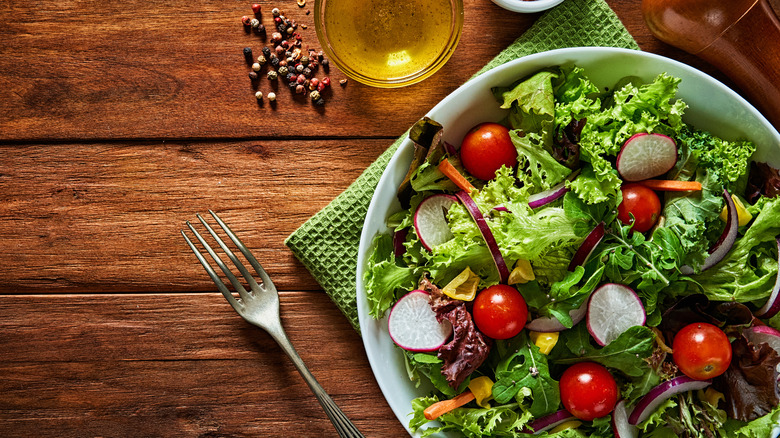 A bowl of salad next to peppercorns and olive oil