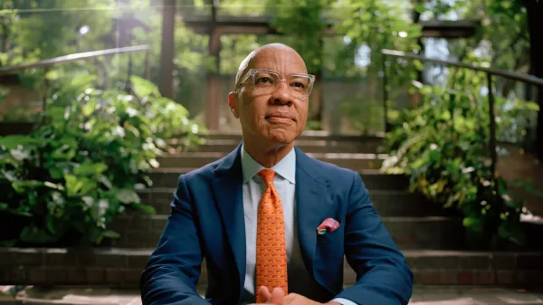 Darren Walker, a bald man with glasses sits confidently in a blue suit, white shirt, and orange patterned tie. He is on outdoor stone steps surrounded by lush greenery, including various plants and trees. Sunlight filters through the leaves, creating a serene atmosphere.