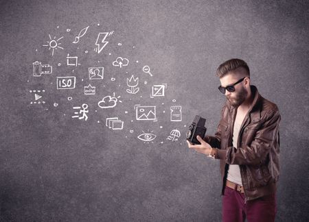 An elegant hipster guy trying to use a vintage camera with camera settings icons drawn on wall, like flash, white balace or ISO value concept