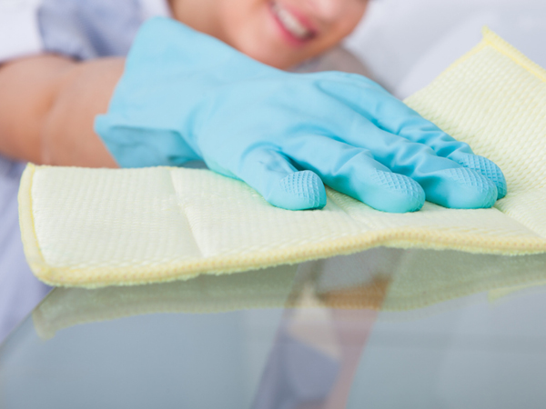 Image shows a woman wearing blue rubber gloves cleaning a glass table with a yellow cloth