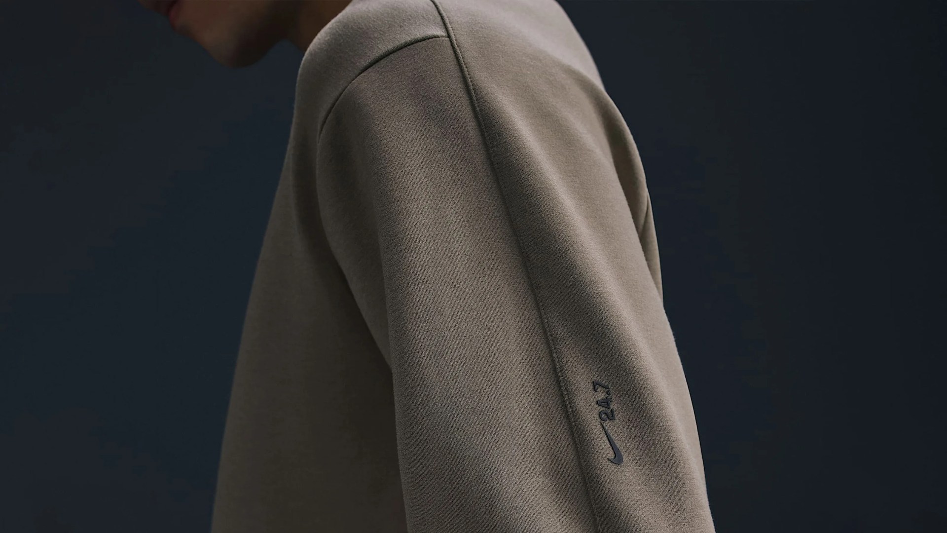 The arm of a Nike sweatshirt in the brands 24.7 collection shown on a model standing in profile against a dark blue background