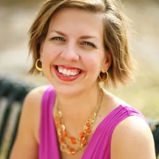 A woman in a purple top and gold necklace smiles on a park bench.