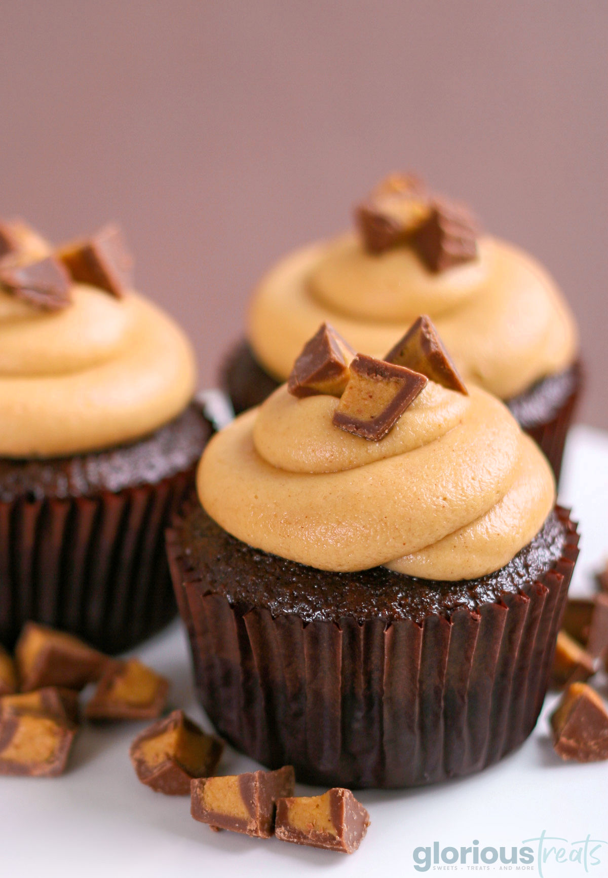 three chocolate cupcakes with peanut butter frosting and chopped Reeses candy on top sitting on a white cake stand.