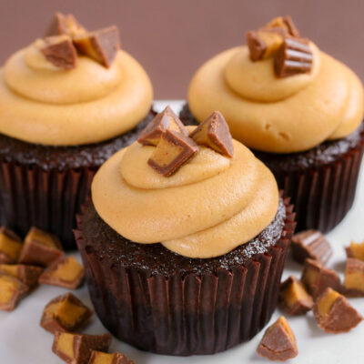 three chocolate cupcakes with swirled peanut butter frosting and chopped Reeses candy on top sitting on a white cake stand.