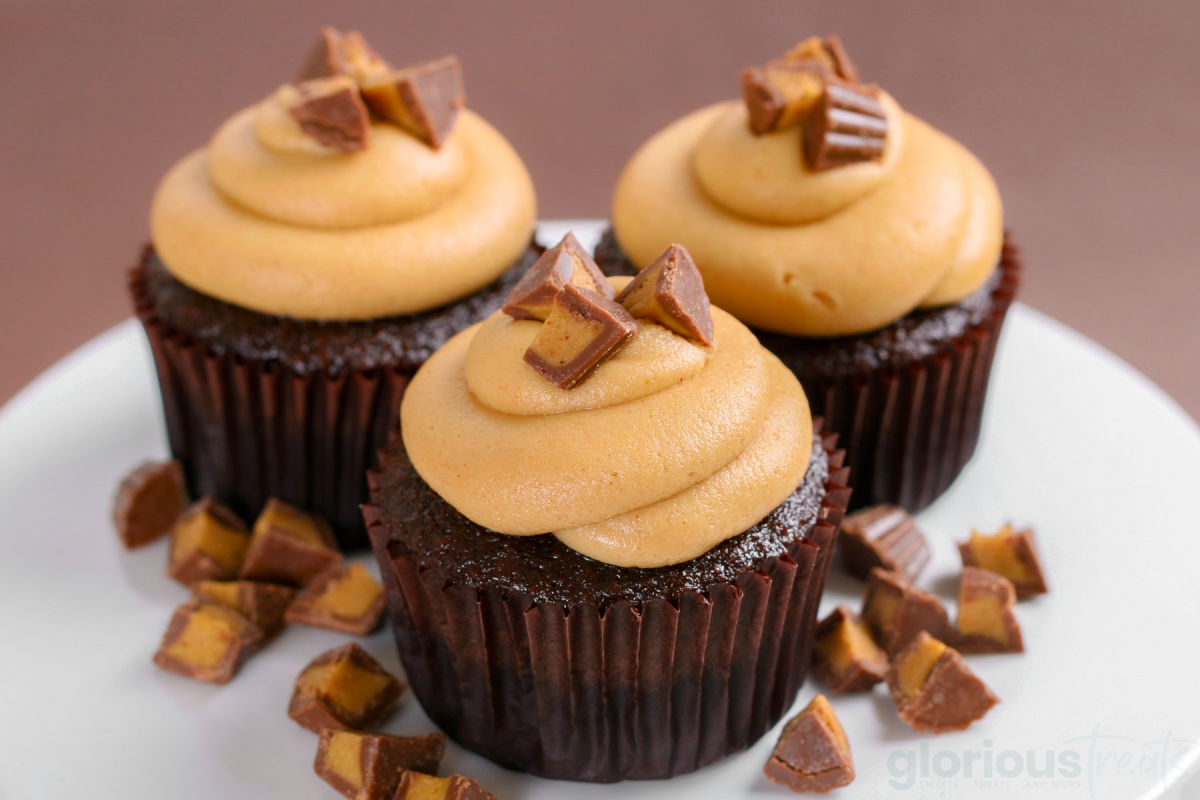 three chocolate cupcakes with swirled peanut butter frosting and chopped Reeses candy on top sitting on a white cake stand.