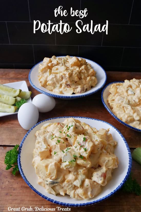 A wood surface with three white bowls filled with potato salad, with a small white plate with pickle spears, and two hard boiled eggs, and the title of the recipe in text at the top.