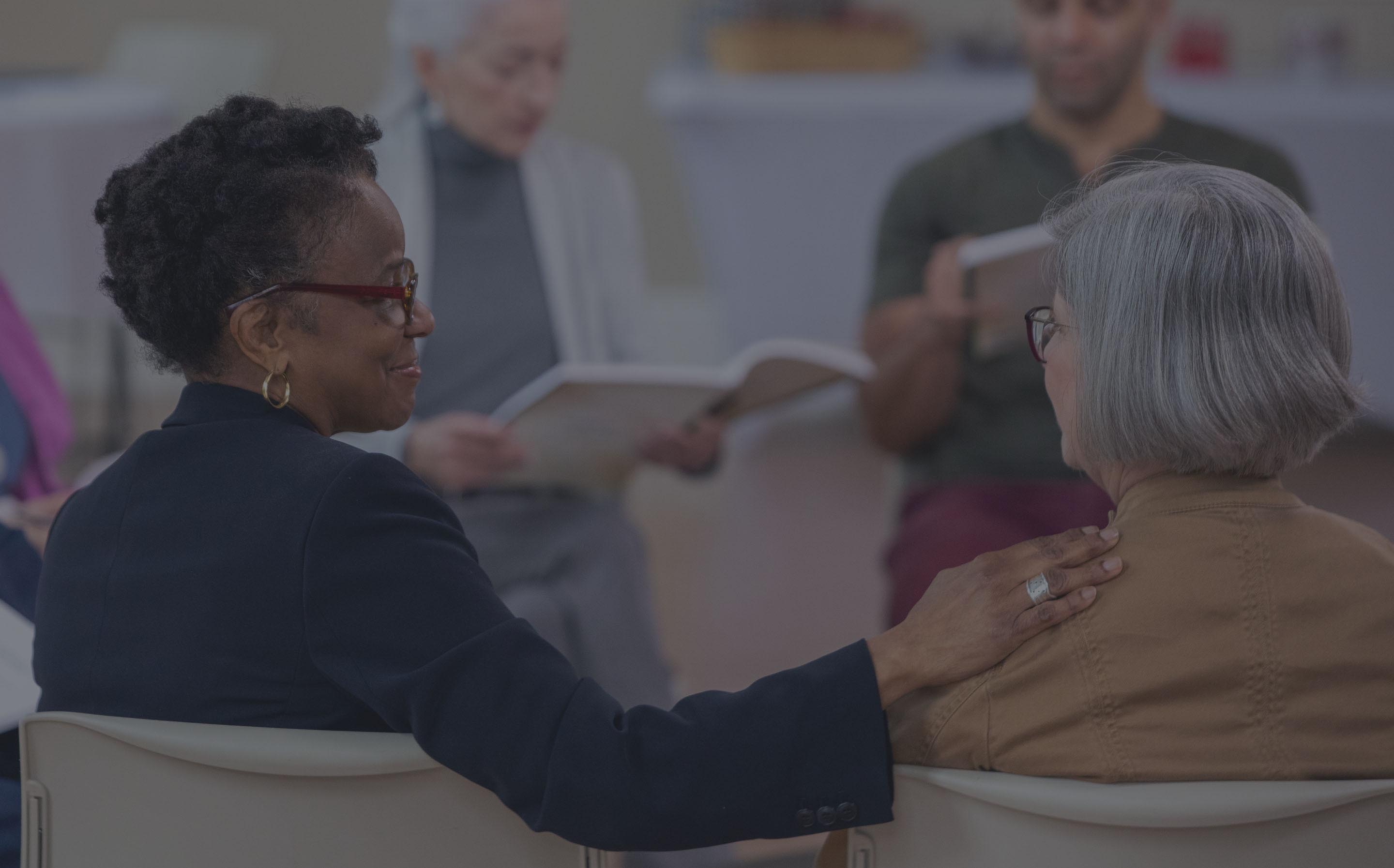 A diverse group of people sit together at a GriefShare support group. They are holding workbooks and warmly interacting.