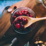 A jar of honey fermented cranberries with a wooden spoon lifting some out, top view. On a dark wood surface.