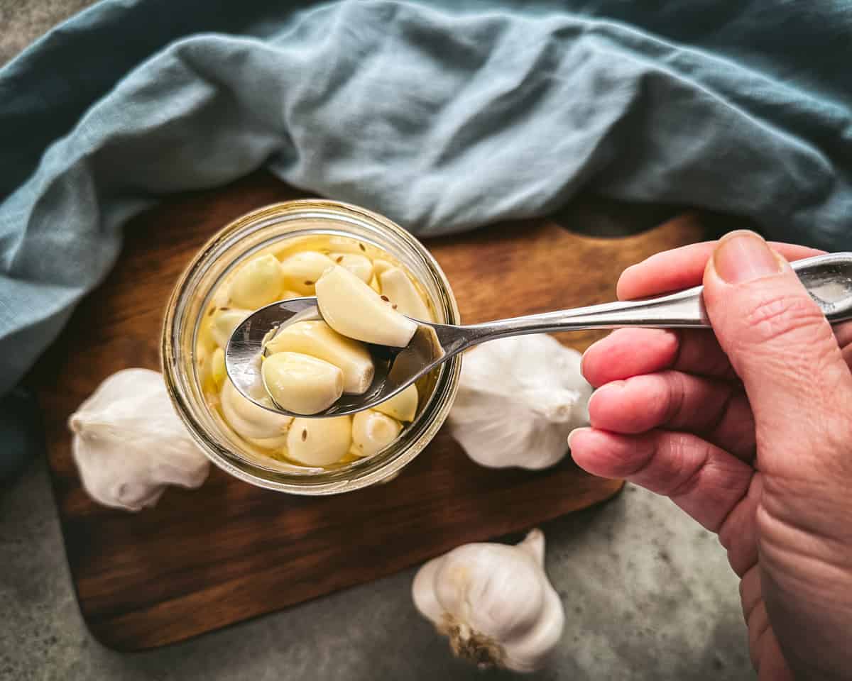 A jar of pickled garlic with a spoon lifting some up, with a background of a blue cloth, a wood cutting board, and garlic heads. Top view.